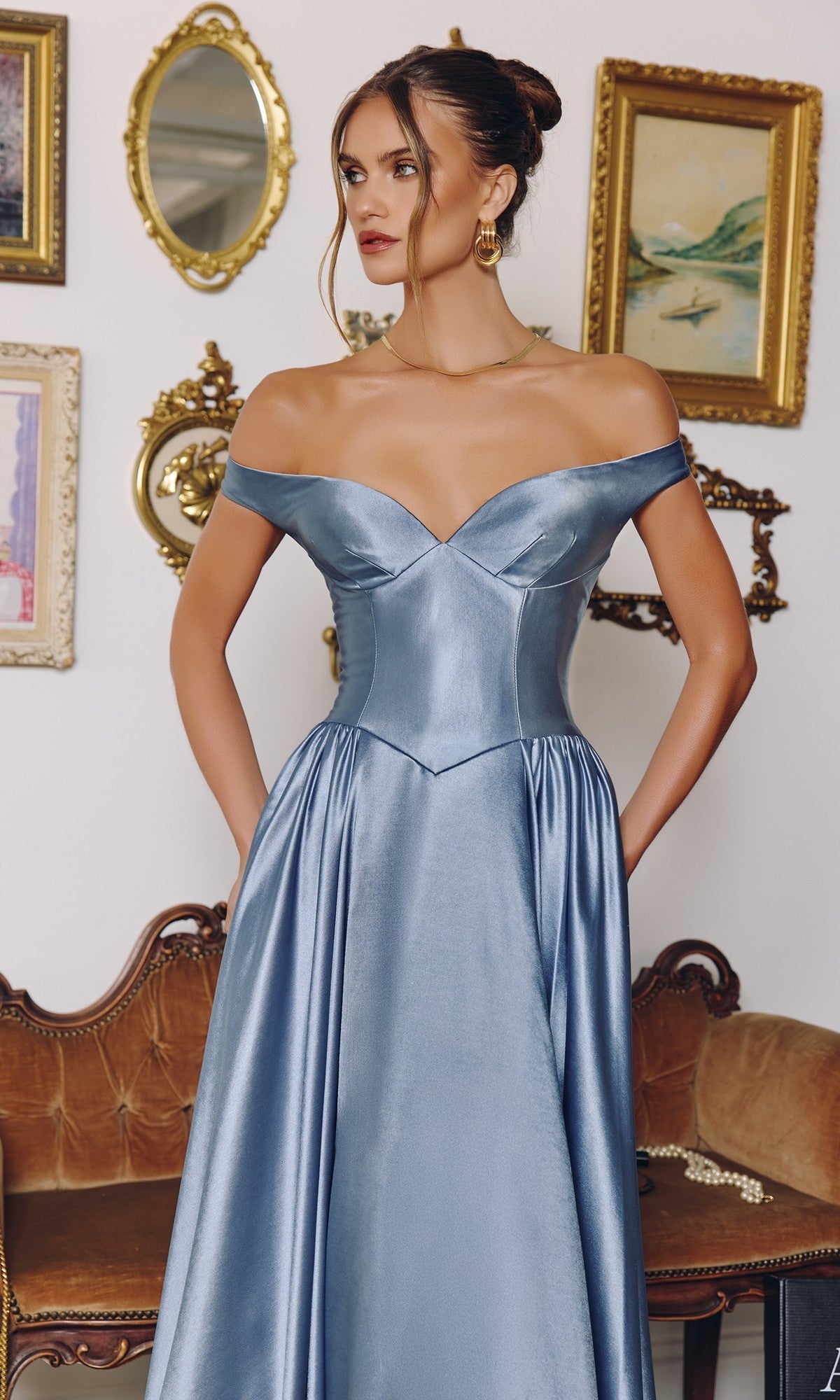 A woman stands before a vintage-style wall and brown sofa, wearing the Amoris T821 long prom dress—an off-the-shoulder, satin blue gown with pockets and an elegant A-line silhouette.